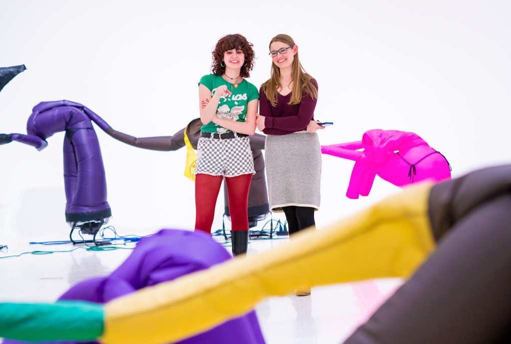 Two adults smile at point at colorful inflatible sculptures that resemble figures in a large white art gallery.