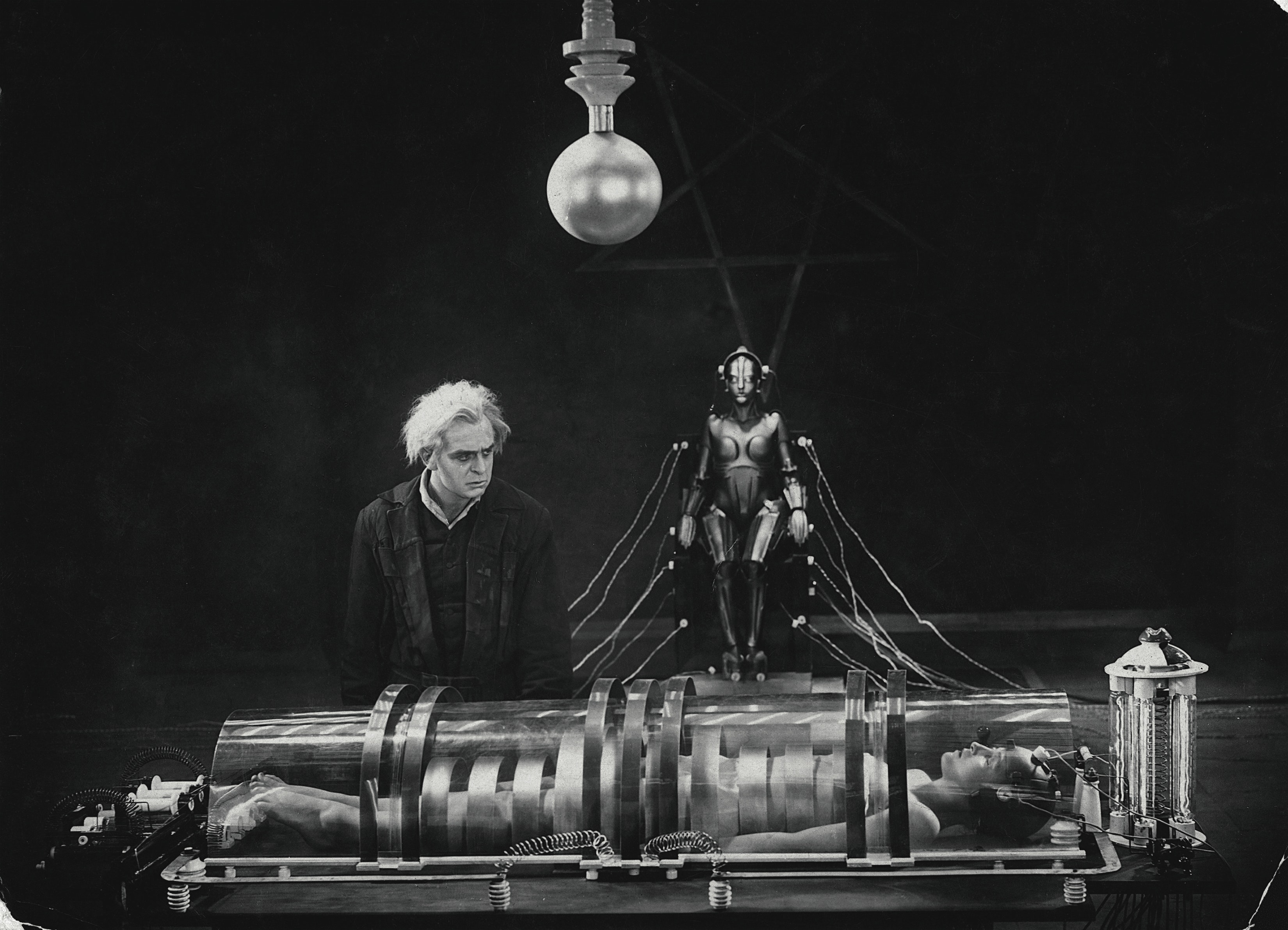 Black and white image of male scientist looking down at woman in chamber connected to a female robot in background