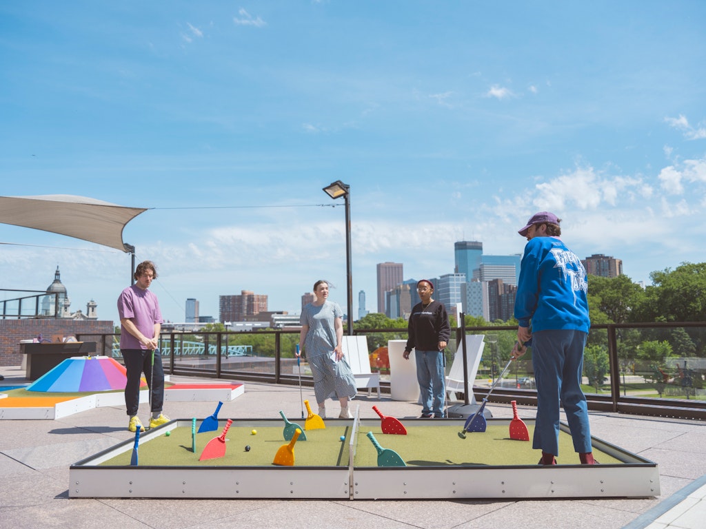 Locally Uploaded Image This image was uploaded directly to AltText AI for processing. Four people play mini-golf on a rooftop with a city skyline in the background under a blue sky.