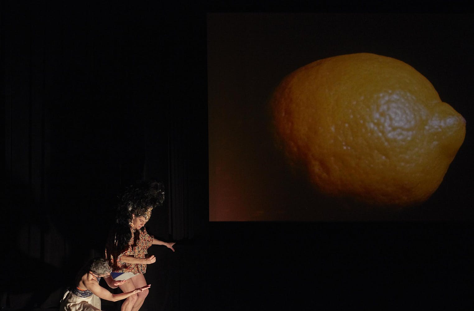 Two dancers perform in front of a large screen with a film of a lemon.