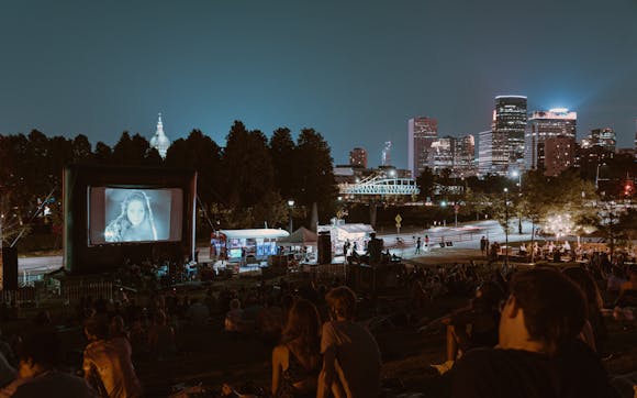 Crowds of people on pinic blankets on a hillside next to a museum watch a film projected on a large outdoor screen with a band playing in front of it and the Minneapolis skyline in the background.