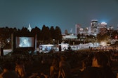 Crowds of people on pinic blankets on a hillside next to a museum watch a film projected on a large outdoor screen with a band playing in front of it and the Minneapolis skyline in the background.