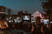 A crowd of people sitting in a park, facing an outdoor screen playing a movie during twilight.