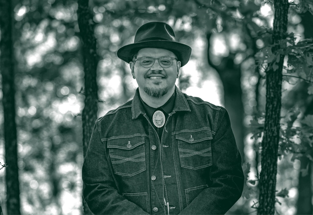 An adult with medium brown skin stands looking at the viewer in a forest. They are wearing a brown fedora and jean jacket.