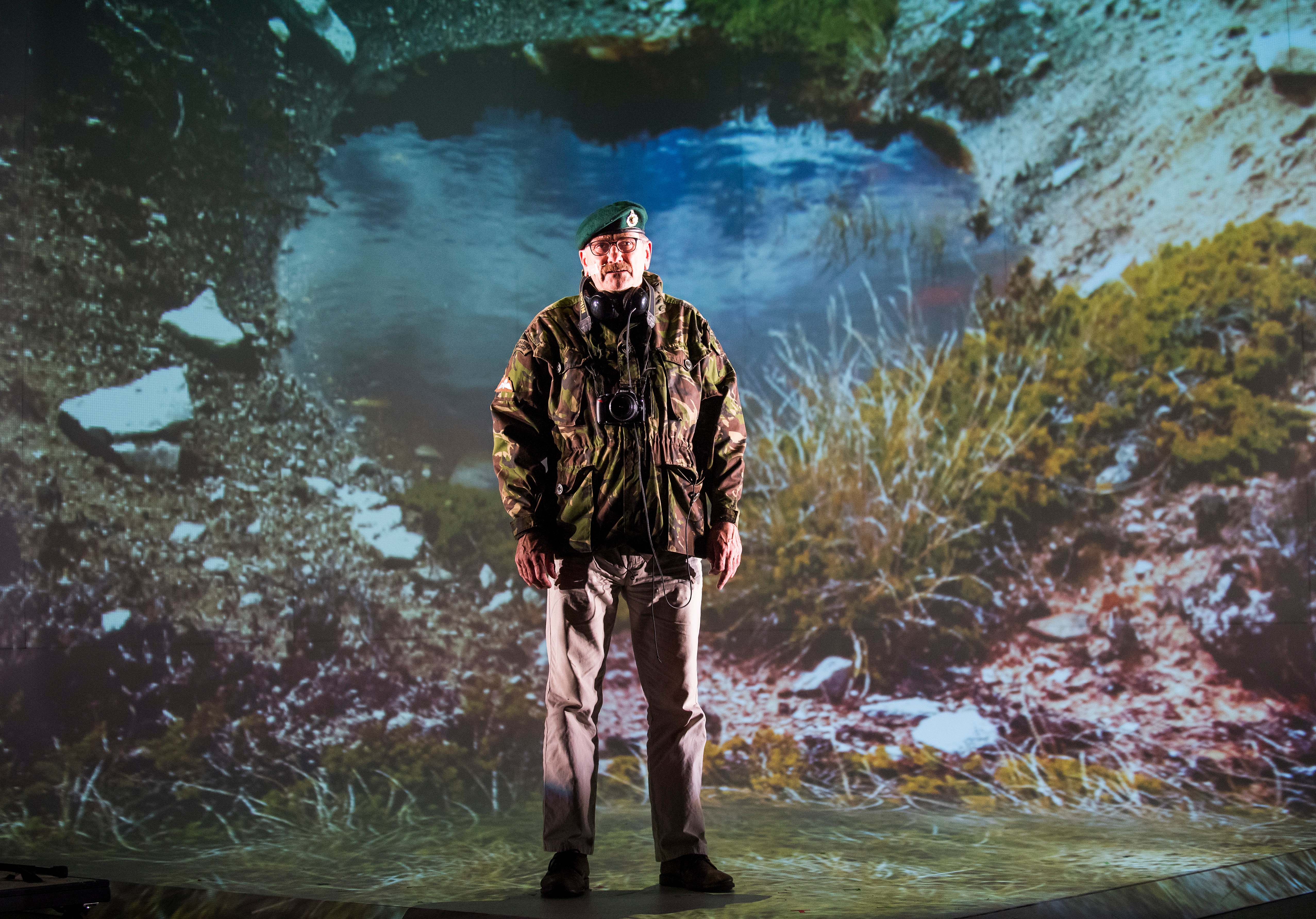 Image from a performance of Lola Arias: Minefield. Man in a military uniform stands in front of a projection of a crater.