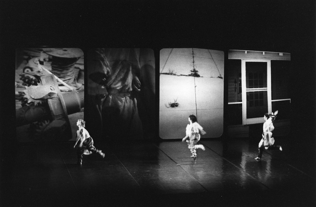 Three dancers in a dramatic stage setting with four large, abstract screens in the background displaying various images.