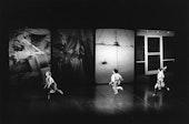 Three dancers in a dramatic stage setting with four large, abstract screens in the background displaying various images.