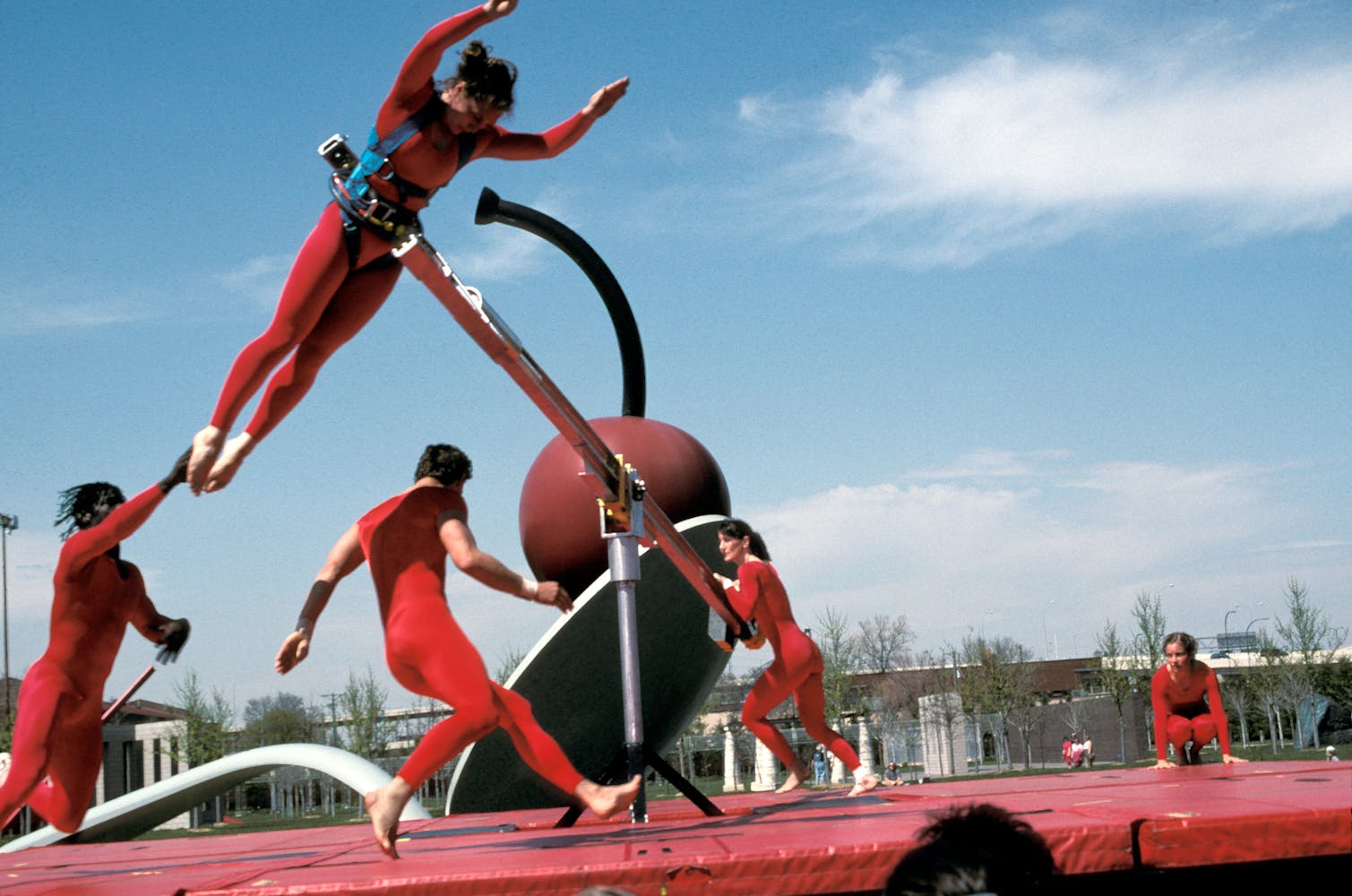 Dancers in red use long poles to jump through the air outside.