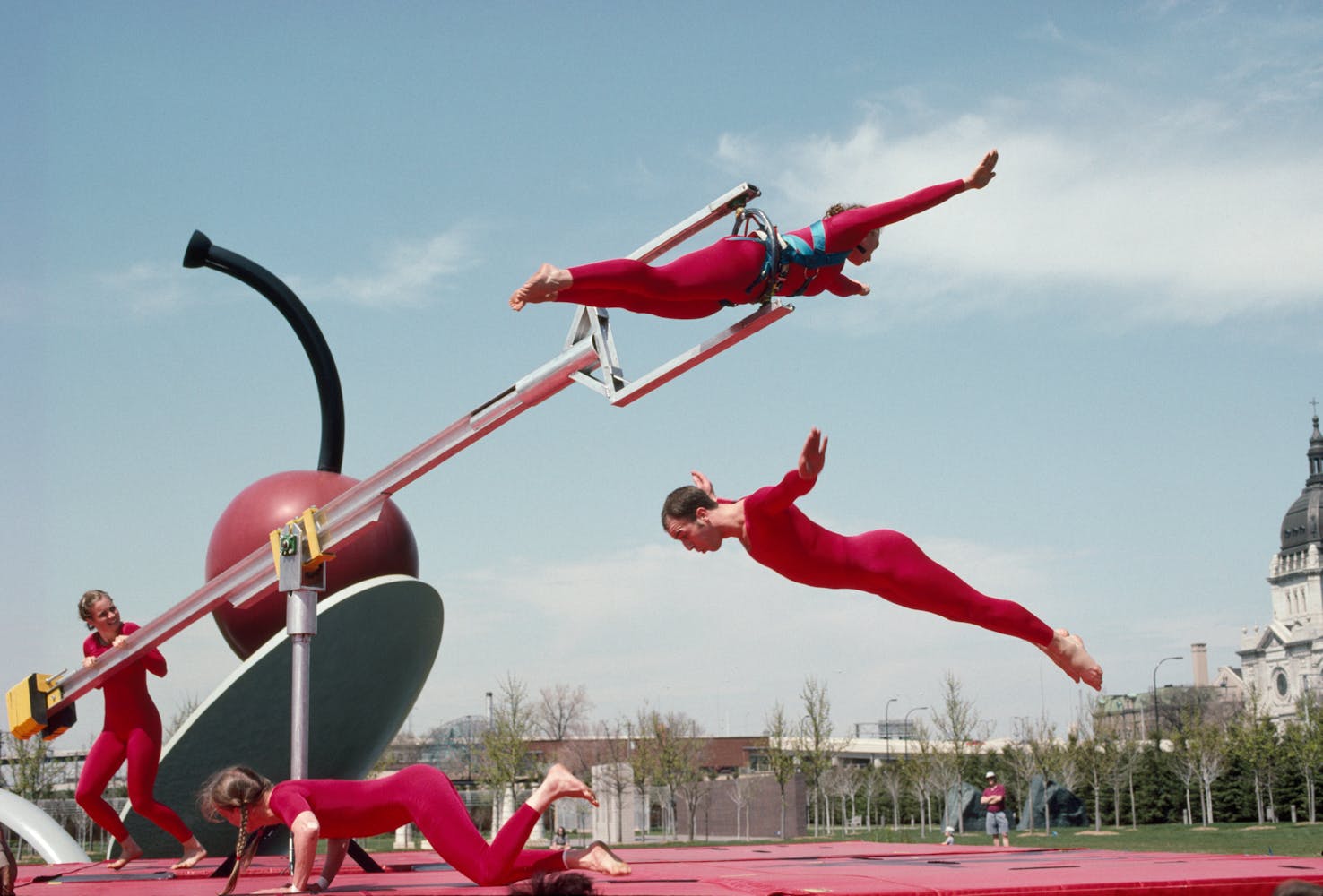 Dancers in red use long poles to jump through the air outside.