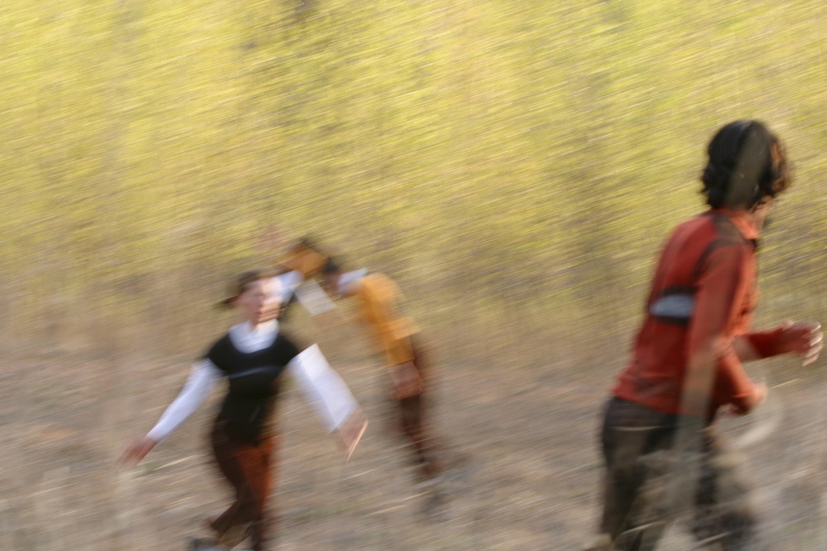 Four dancers run through a crop field, their paths intersecting. The image is blurred due to the dancers’ movement.