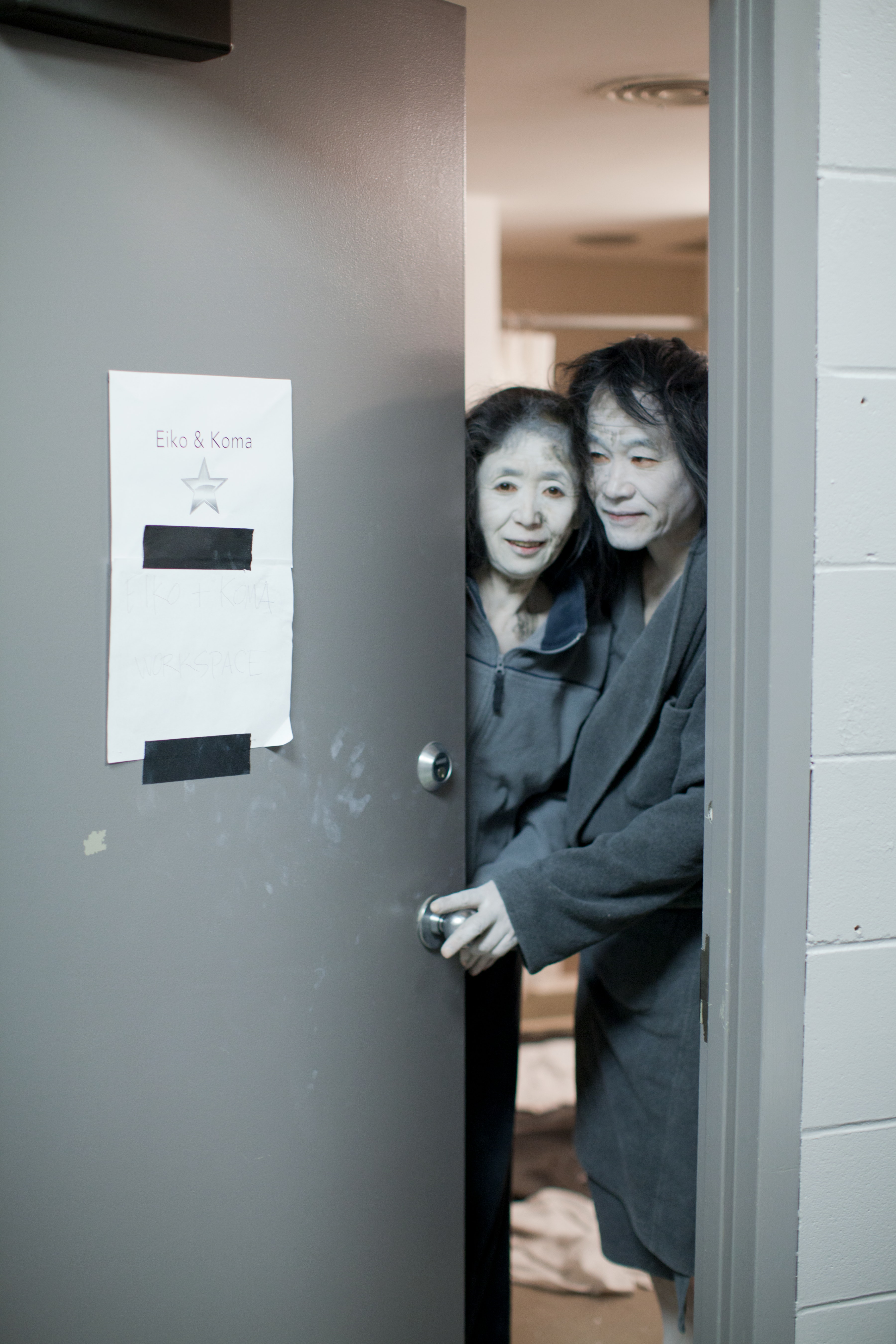 Two people in robes stand in a doorway, looking out from a room with a sign that reads Eiko & Koma.