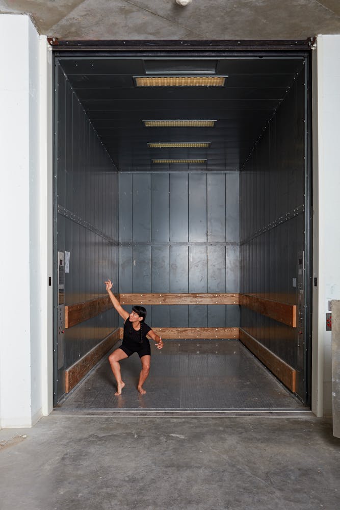 An adult wearing a black dancing leotard croutches and holds their hand up in an empty frieght elevator.