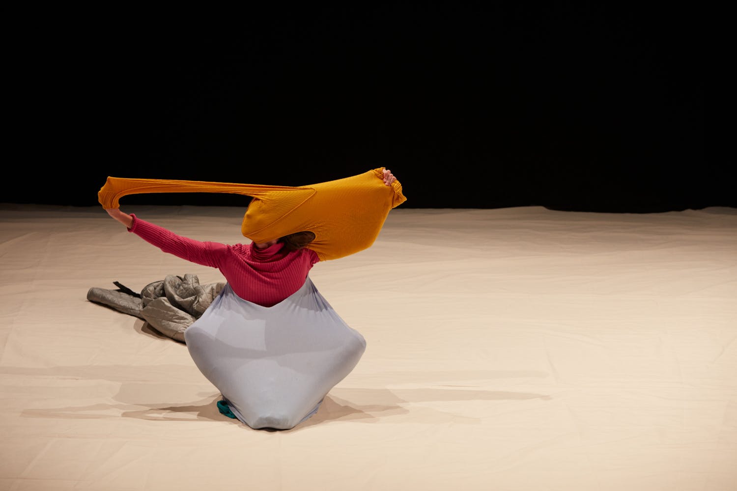 A dancer with white skin and brown hair sits on a stage with gray fabric stretching around their torso and legs. They wear a rose-pink turtle neck and pull a yellow sweater over their head, obscuring their face.