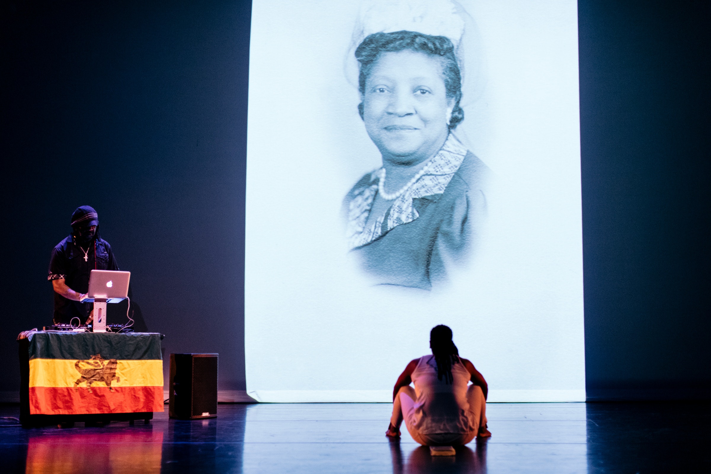 A woman sits on stage with her back to the viewer and watches projected images of old photographs of women while a man DJ's from a table nearbby.