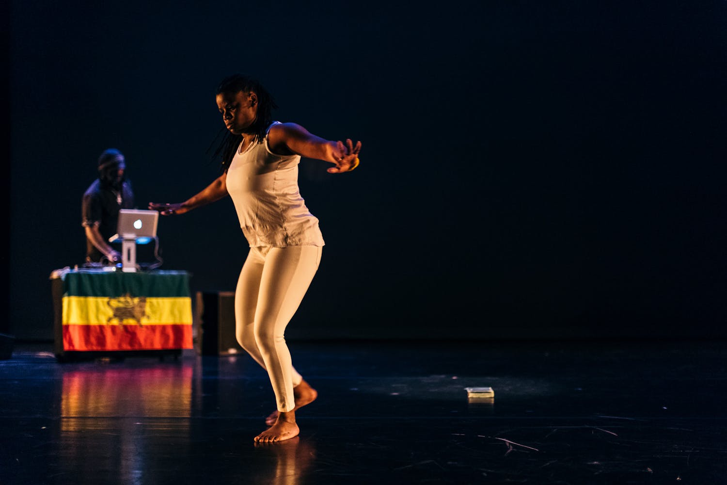 A womand dances on stage in front of a book while a man DJ's from a table on the stage nearby.