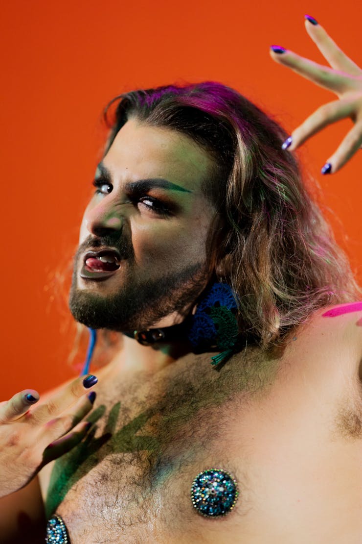 Shirtless dancer wearing bright colored pasties and snarling with hands elevated and red background