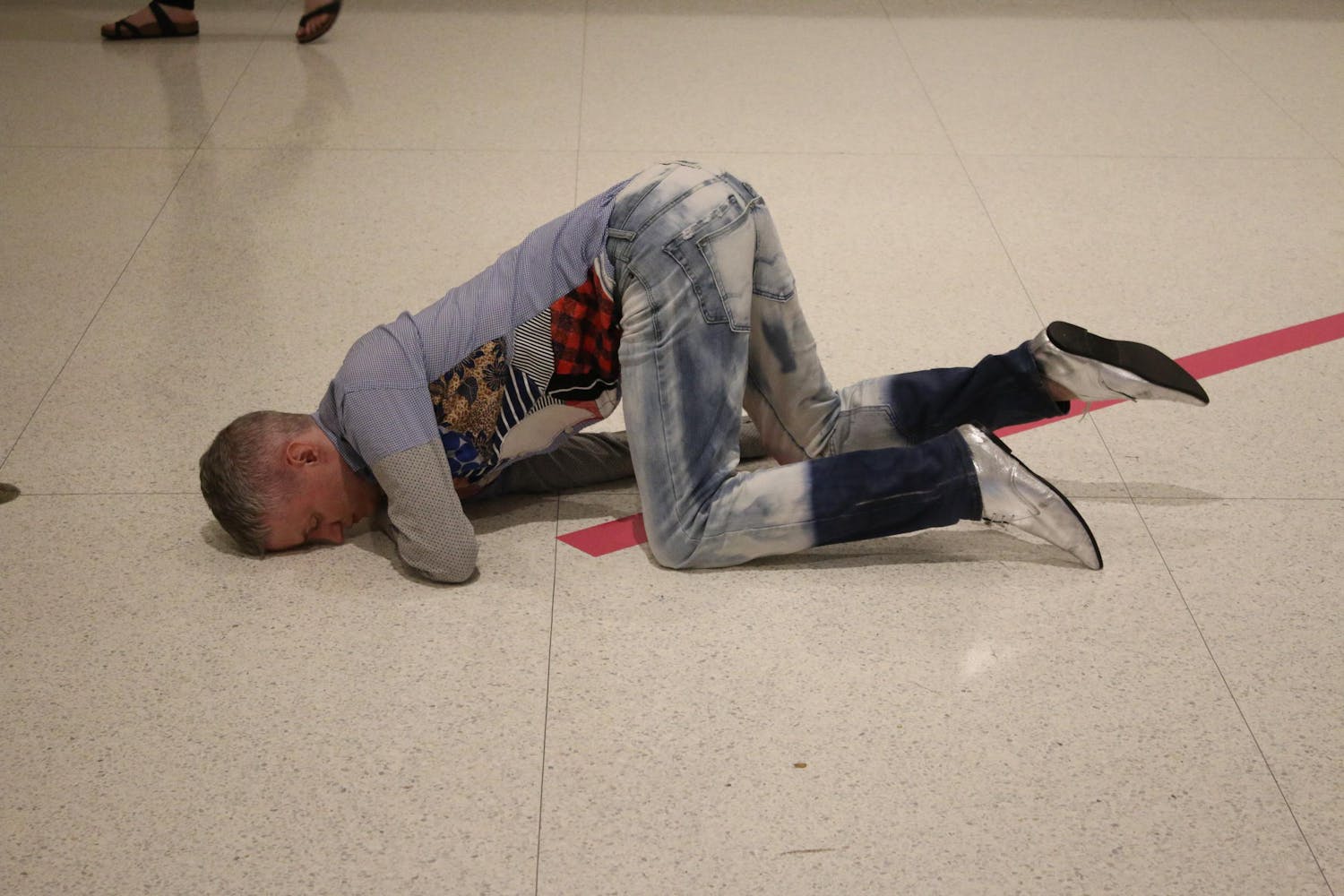 A dancer with white skin and short grey hair kneels over a thick, pink line taped onto a tiled floor. They bend over, balancing on both forearms with their forehead pressing against the floor.