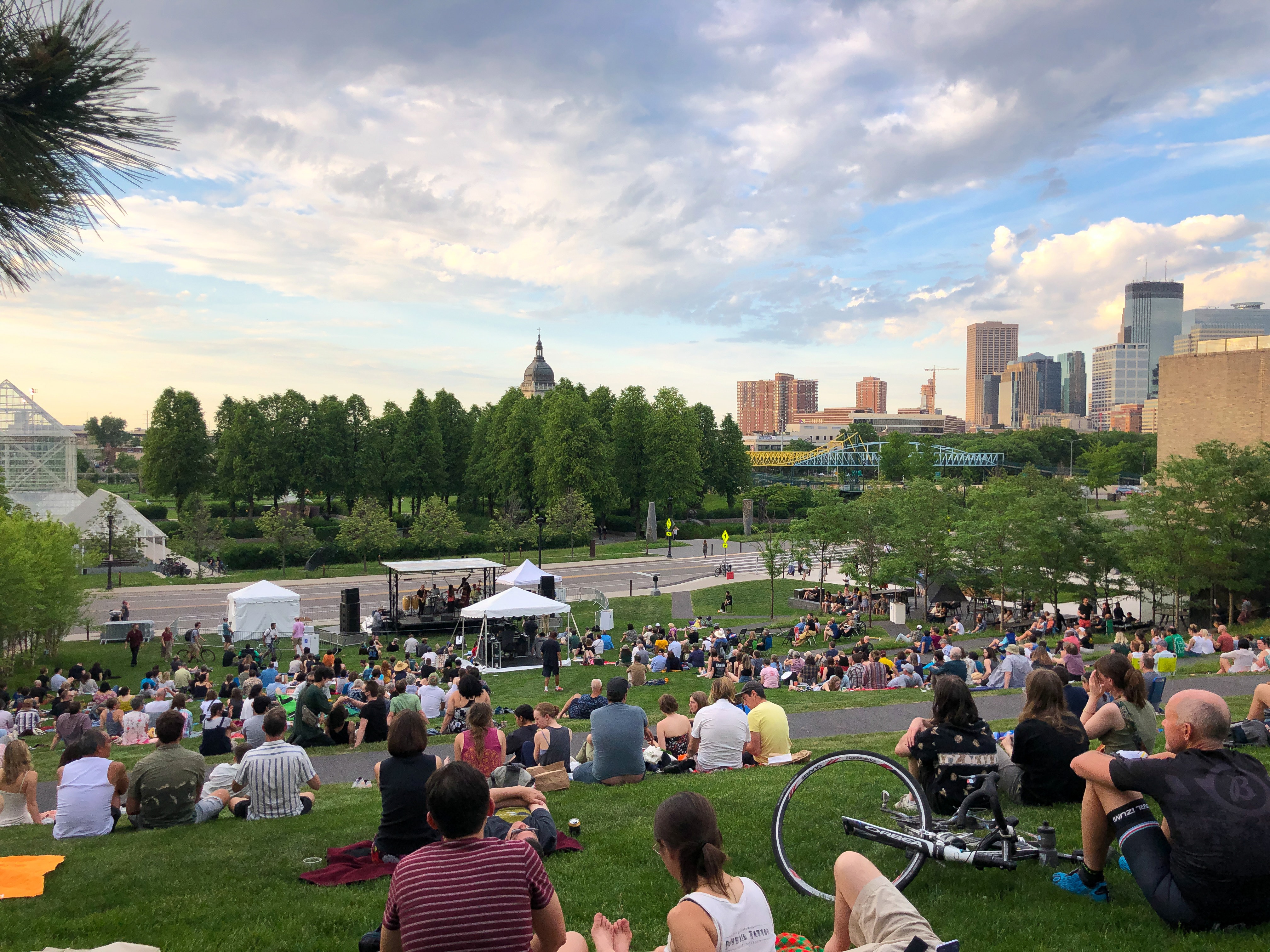Groups of people sit on an outdoor hillside watching a band perform on a stage with a view of the MInneapolis skyline in the background.