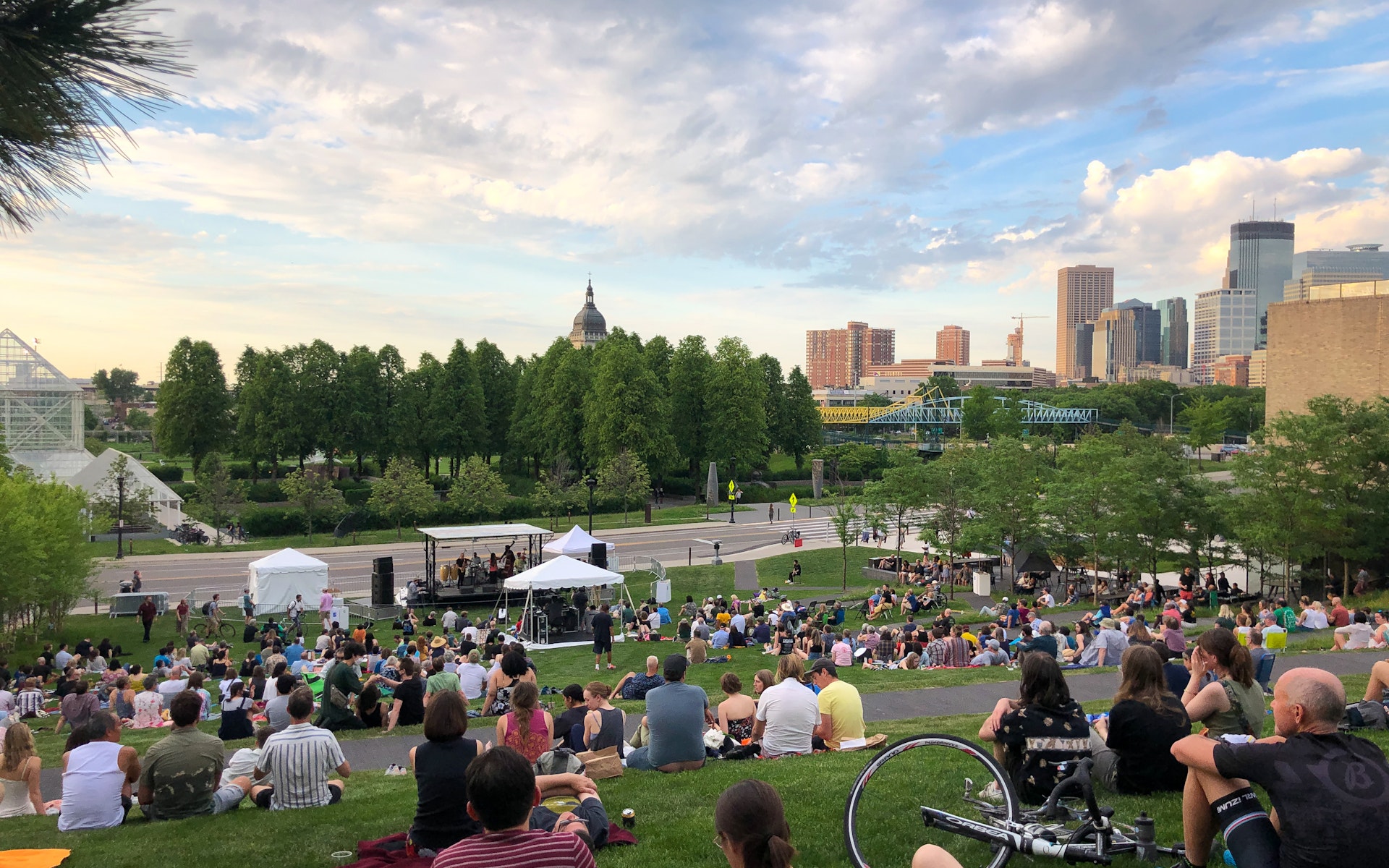 Groups of people sit on an outdoor hillside watching a band perform on a stage with a view of the MInneapolis skyline in the background.