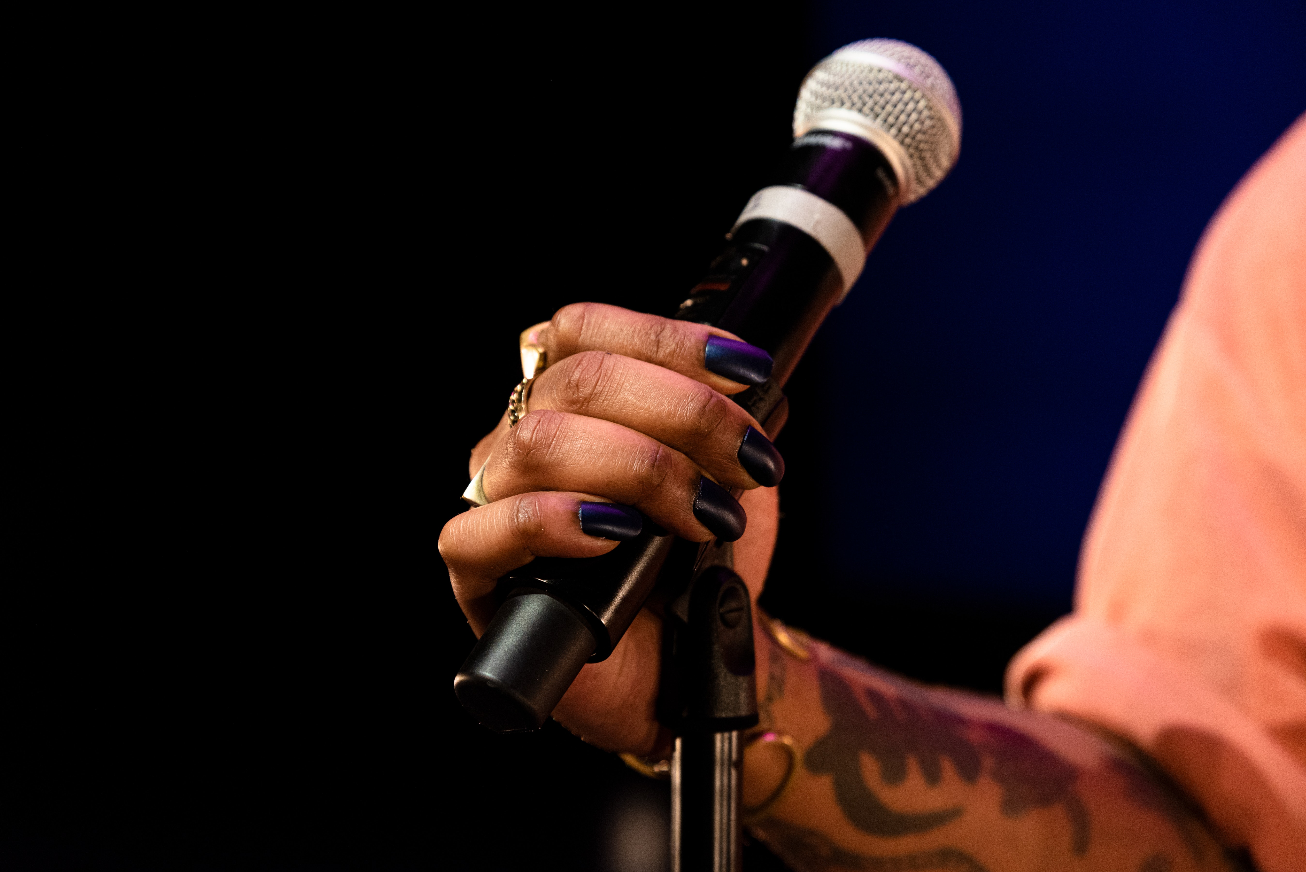 A hand with dark skin and black nailpolish holds a mircophone on a stand.