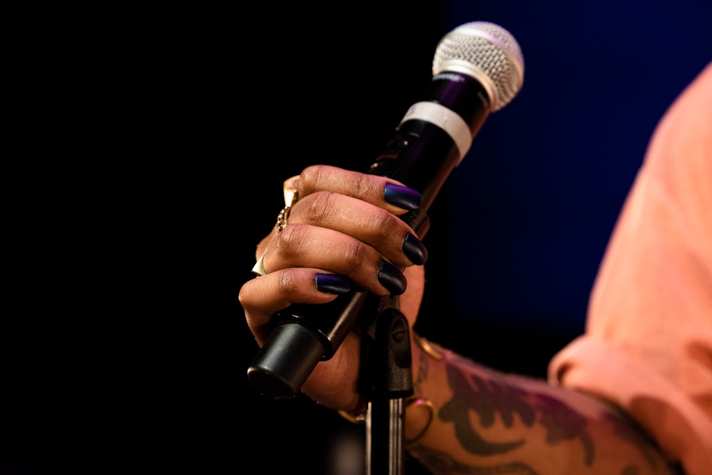 A hand with dark skin and black nailpolish holds a mircophone on a stand.