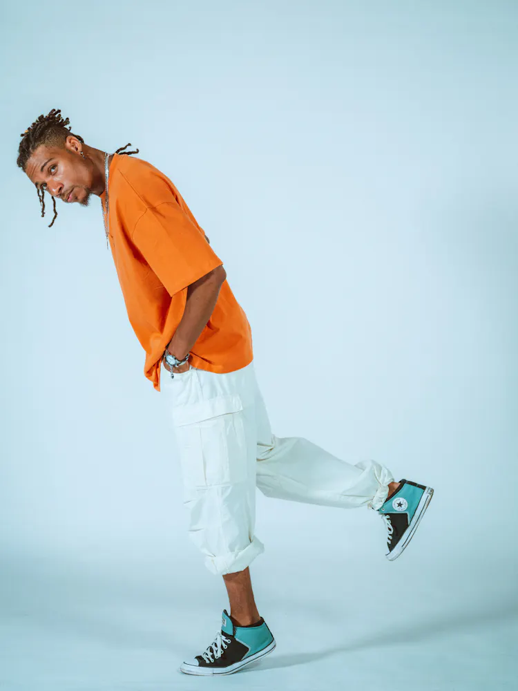 A man with dark skin wearing a t-shirt leans forward on one foot and looks at the viewer.