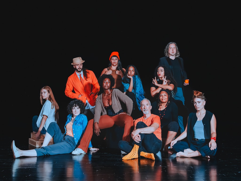 A group of 13 people are arranged on a stage with a black floor and a black curtain behind them.