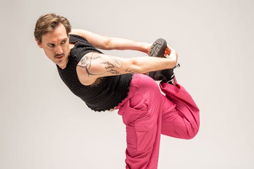 A dancer with light skin and a mustache holds his right leg behind his back.