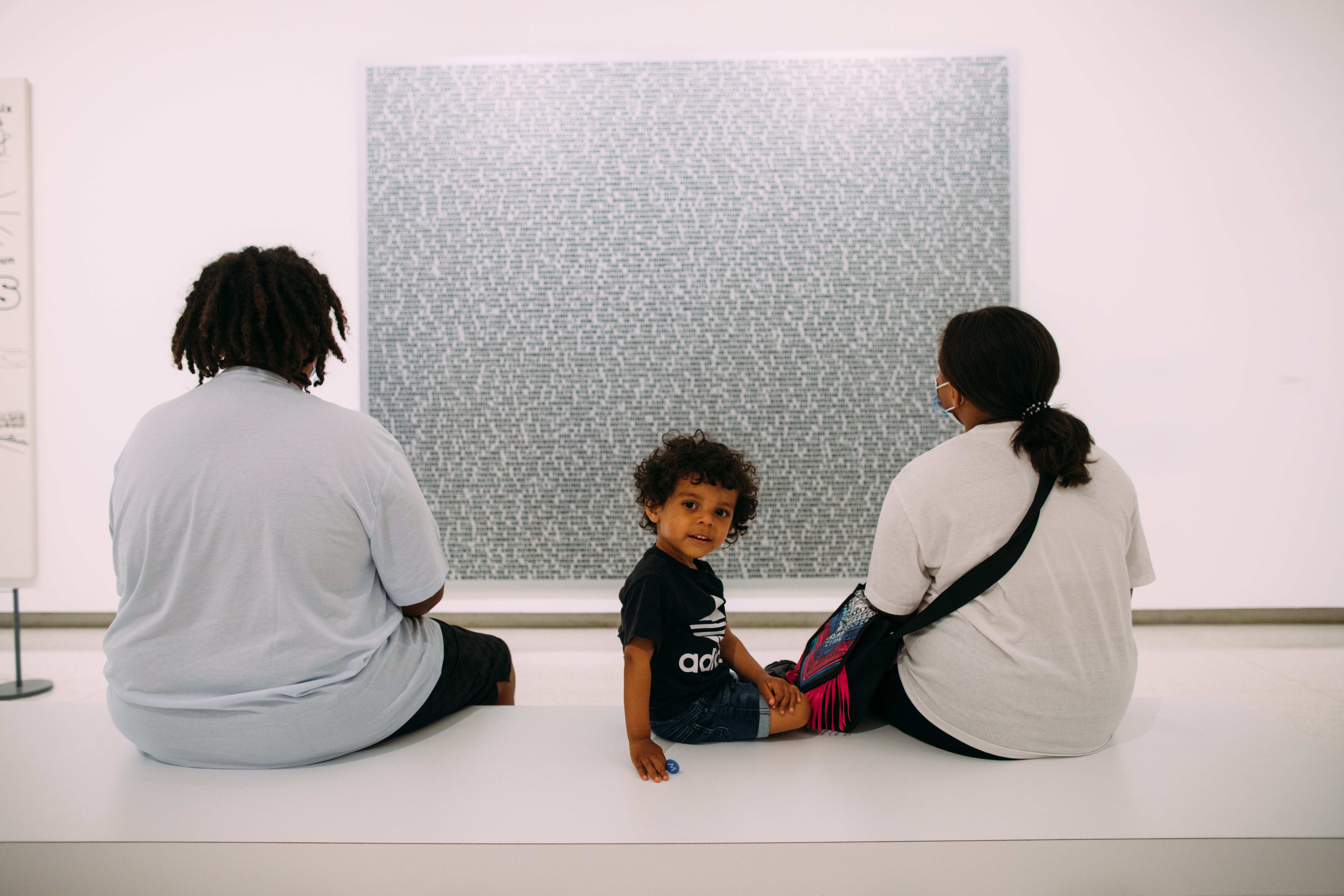 Two adults sit on a museum bench with their backs to the viewer while looking at a painting. Next to them a child turns around and looks at the viewer.