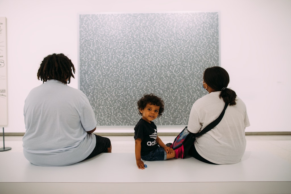 Two adults sit on a museum bench with their backs to the viewer while looking at a painting. Next to them a child turns around and looks at the viewer.