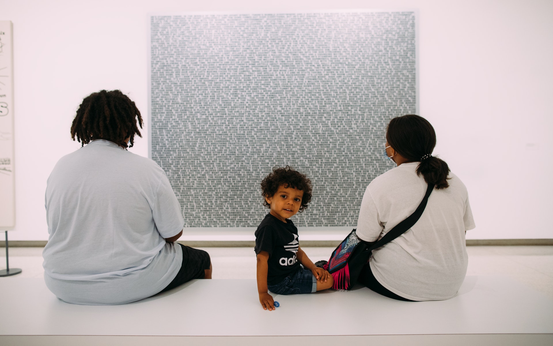 Two adults sit on a museum bench with their backs to the viewer while looking at a painting. Next to them a child turns around and looks at the viewer.