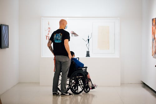One person who is standing and another person who is in a wheelchair looking at art in a gallery, seen from behind