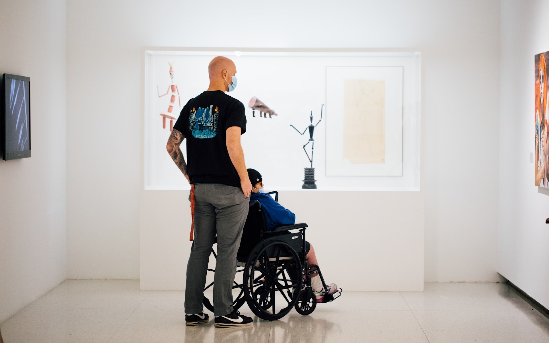 One person who is standing and another person who is in a wheelchair looking at art in a gallery, seen from behind