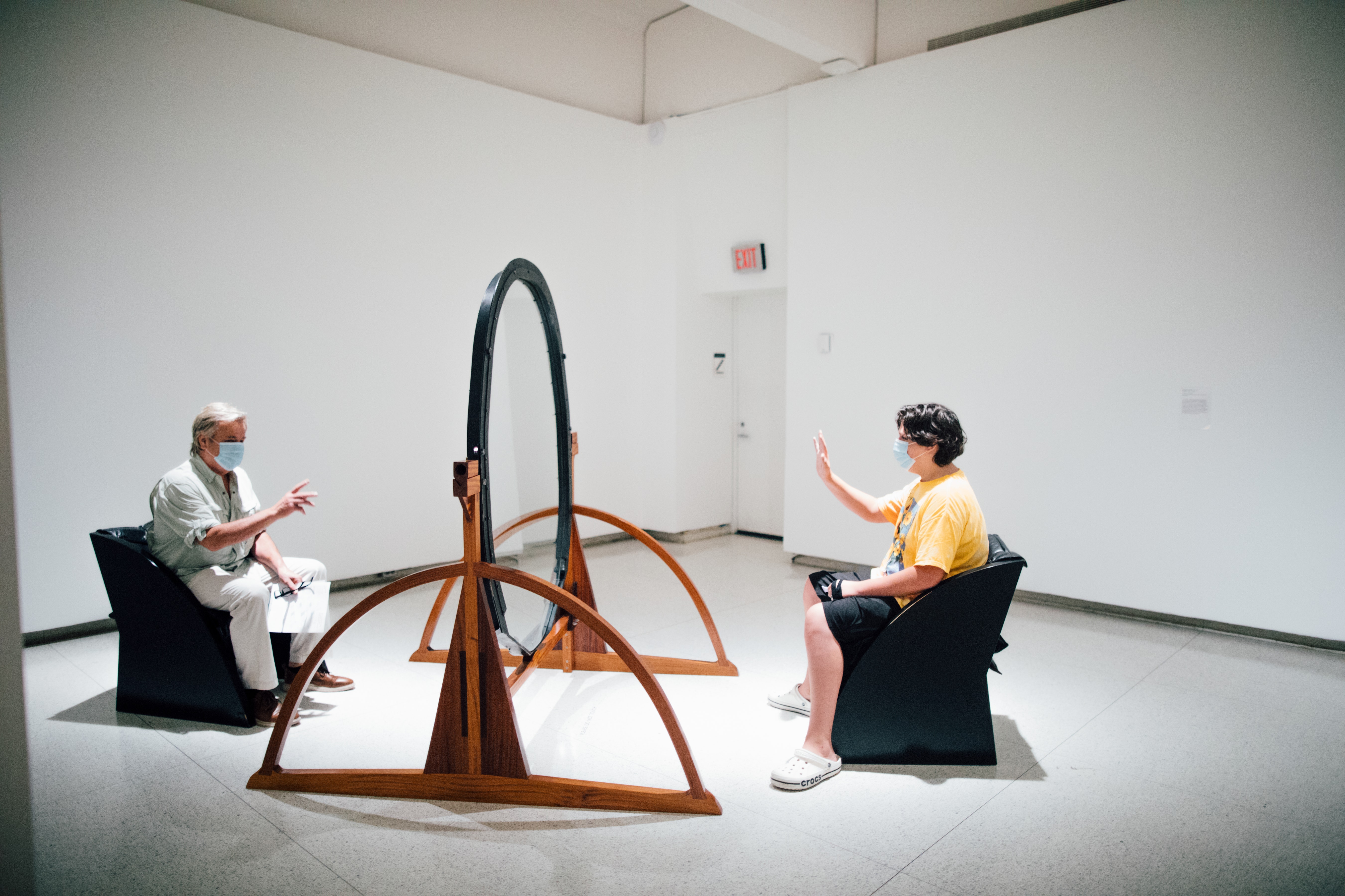 Two people sit in a sculpture consisting of chairs facing each other with glass in between.