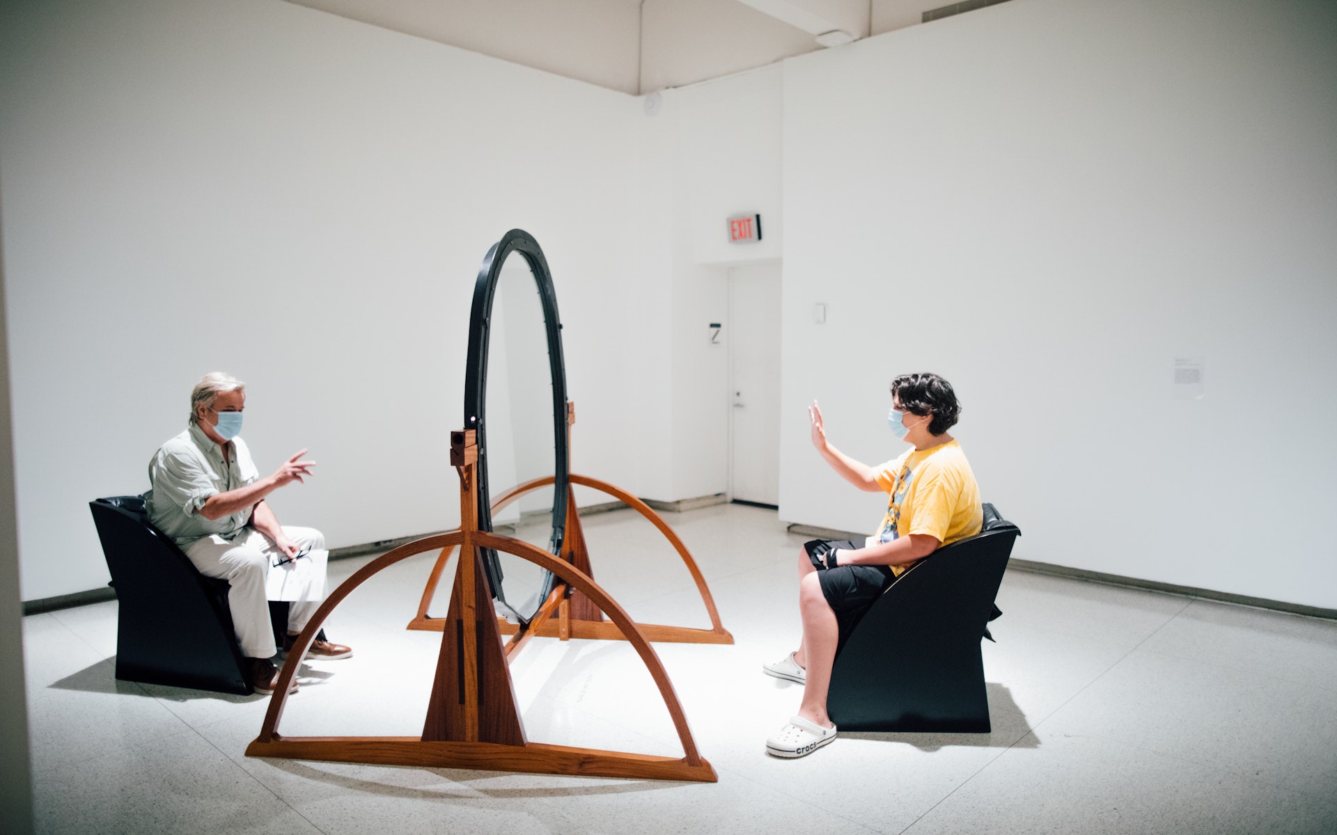 Two people sit in a sculpture consisting of chairs facing eahc other with glass in between.
