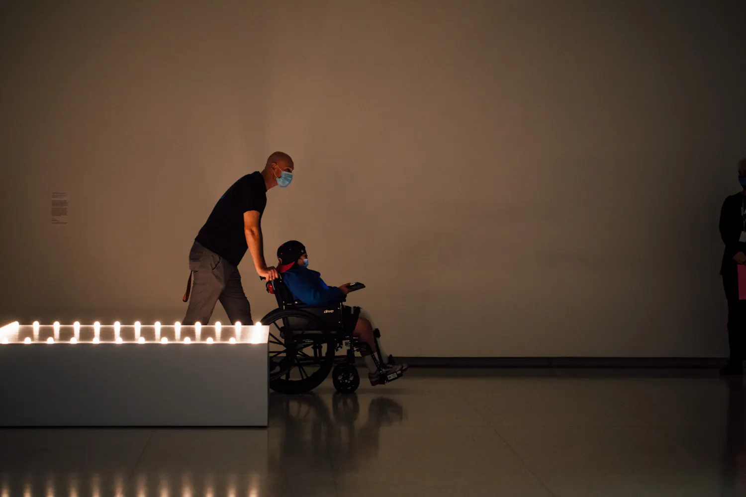 A man pushes a person who uses a wheelchair through a dim gallery.