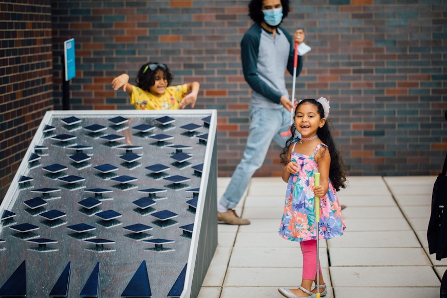 A child smiles while holding a mini gold club next to a large mini-golf hole as an adult in a mask holding a mini golf club and another child look on in the background.