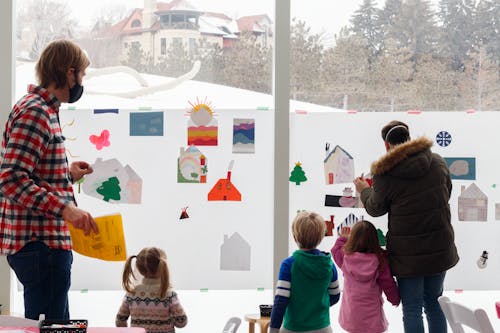 Adults and children adding artwork to a collaborative artwork hung on a window with a snowy hillside visible behind the window