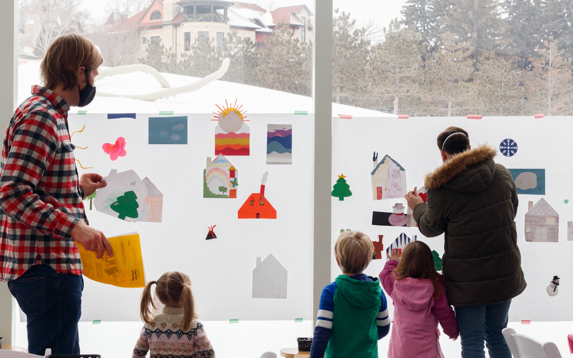 Adults and children adding artwork to a collaborative artwork hung on a window with a snowy hillside visible behind the window