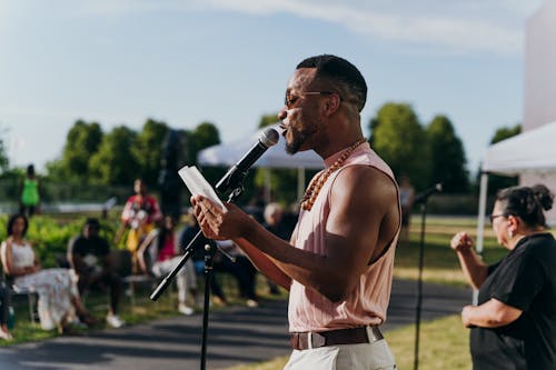 An adult reads into a microphone on a stand from notes they hold while addressing a corwd of people outside on a sunny day.