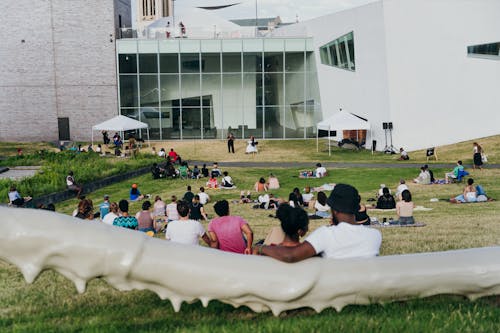 A crowd sits on a lush hillside with large public art sculptures as they listen to adults reading poetry on a stage set up outside of a museum building.