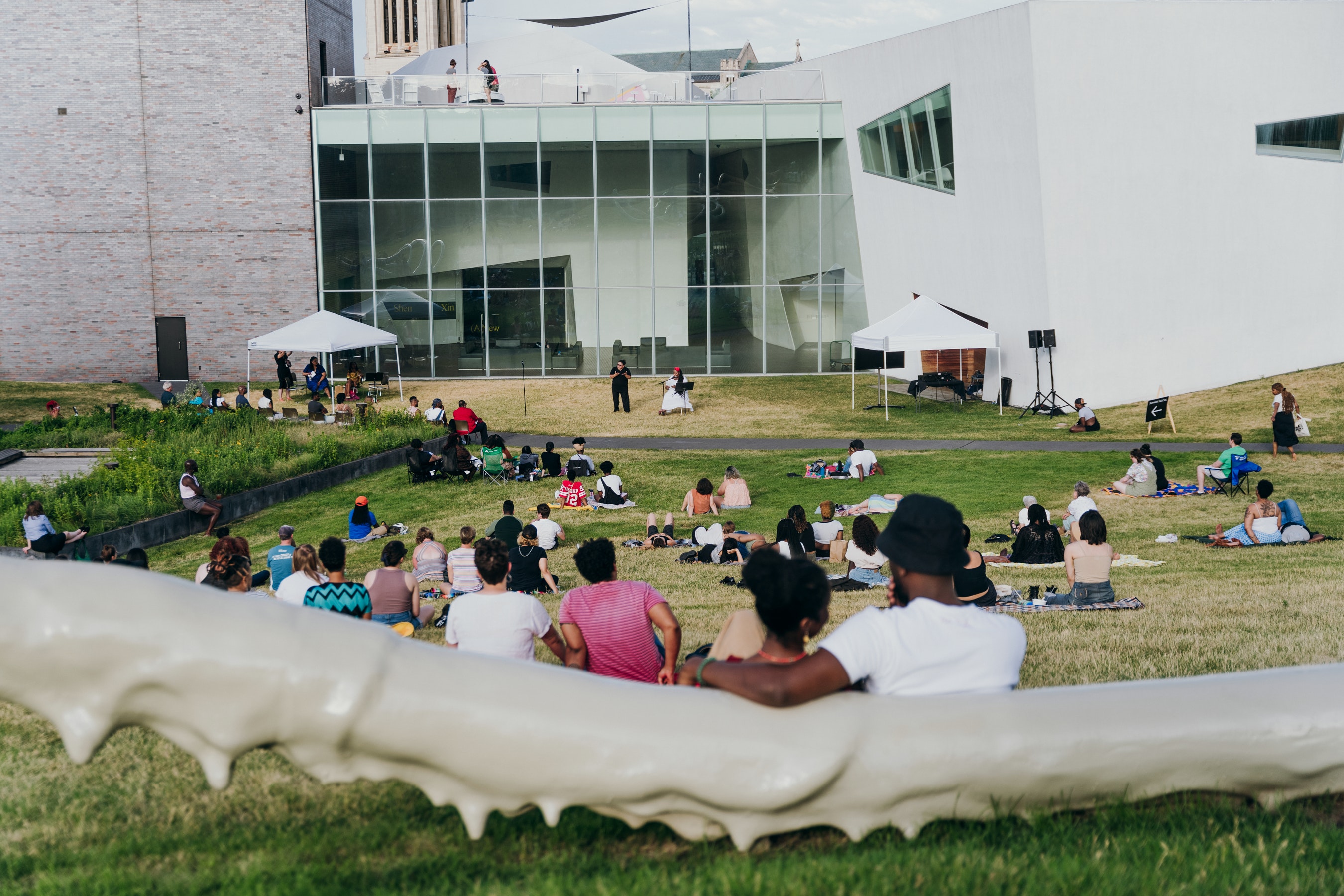Adults sit on a hillside outdoors and lsiten to a person speaking at a microphone