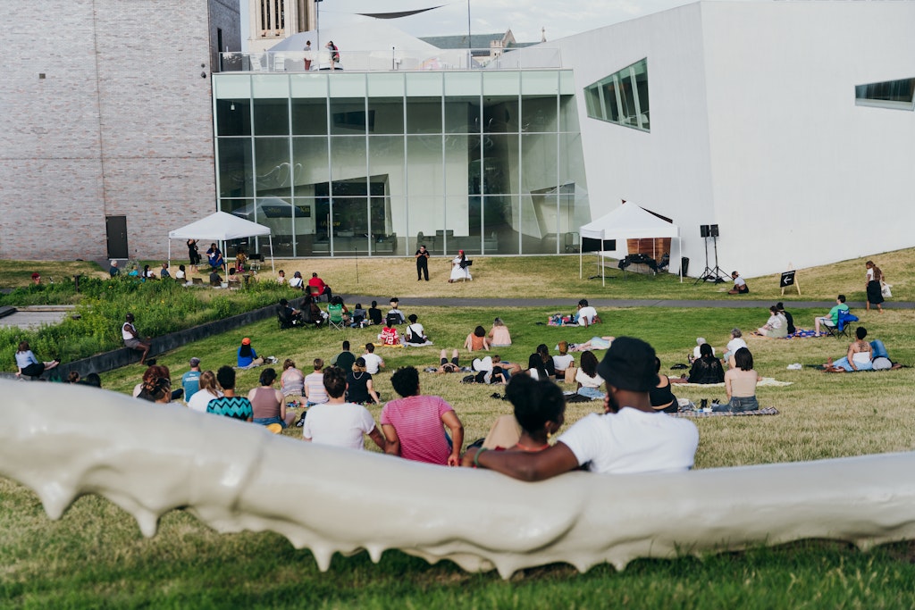 Adults sit on a hillside outdoors and lsiten to a person speaking at a microphone
