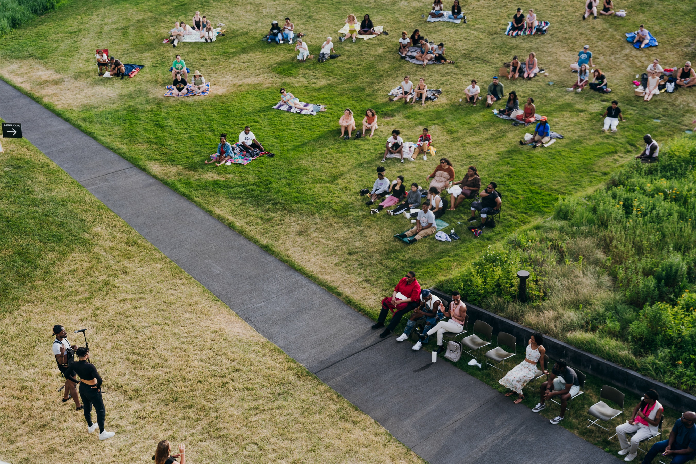 Adults sit on a hillside outdoors and lsiten to a person speaking at a microphone