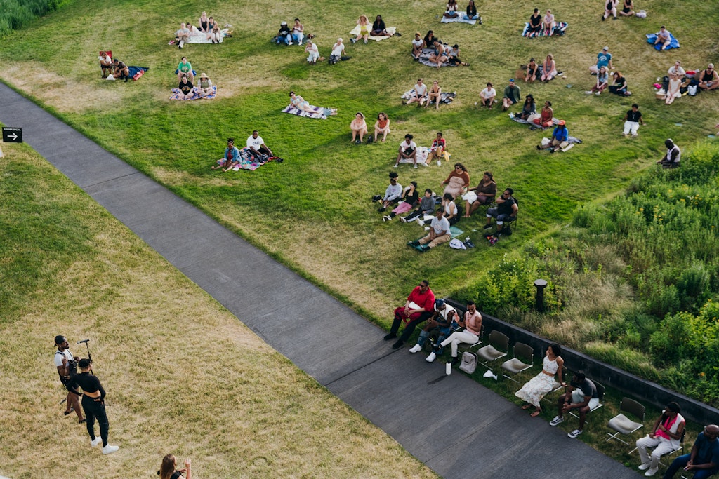 Adults sit on a hillside outdoors and lsiten to a person speaking at a microphone