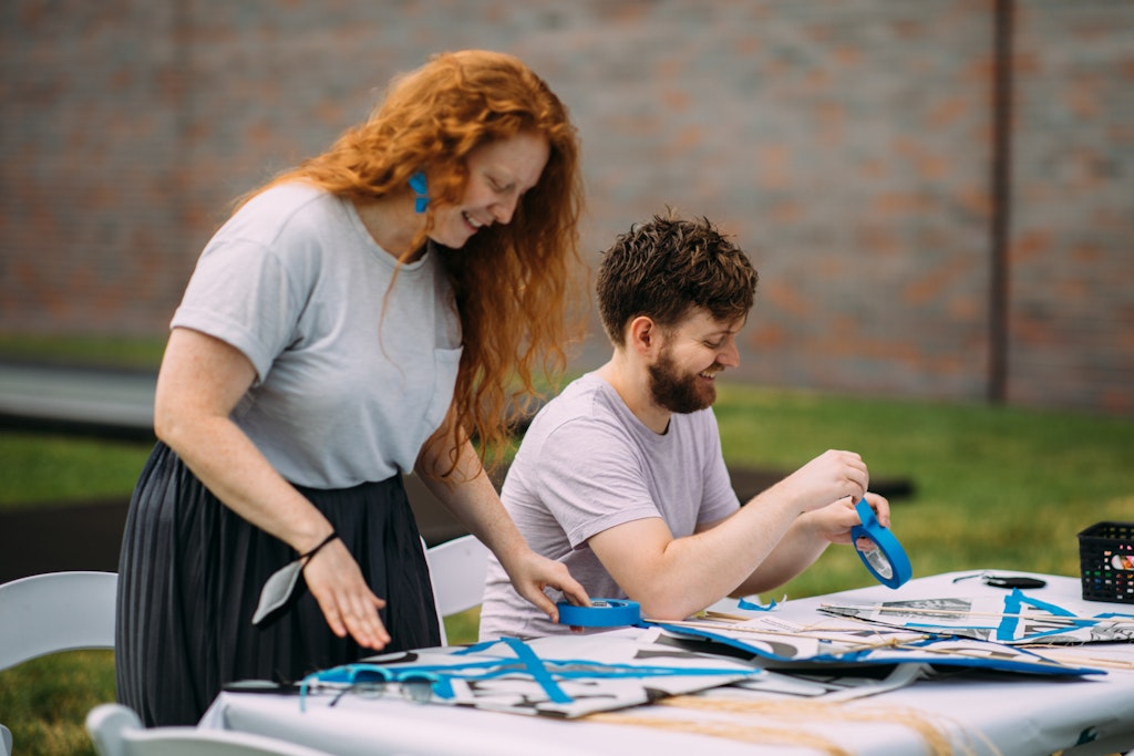 Two people working on arts and crafts with blue tape at a table outdoors.