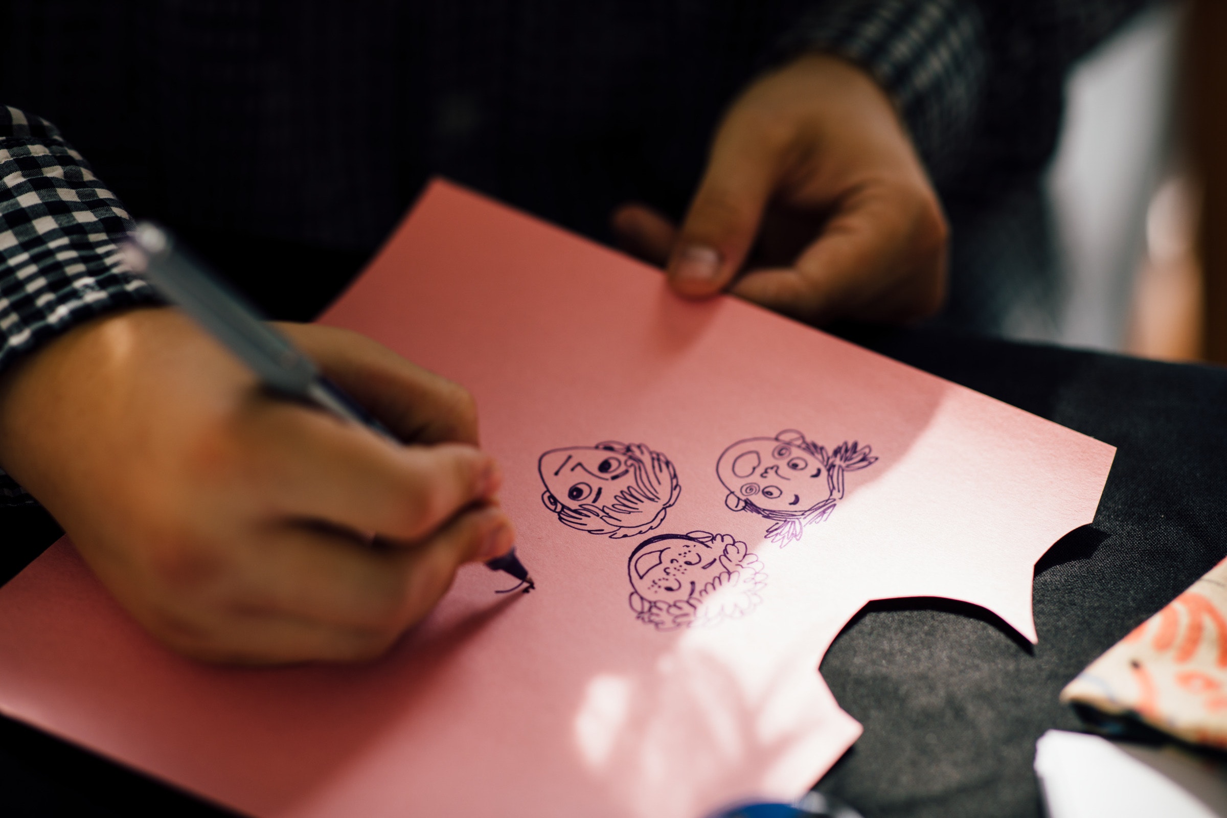 Person drawing cartoon faces with a pen on pink paper, holding it with one hand and sketching with the other.