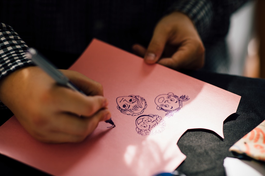 Person drawing cartoon faces with a pen on pink paper, holding it with one hand and sketching with the other.