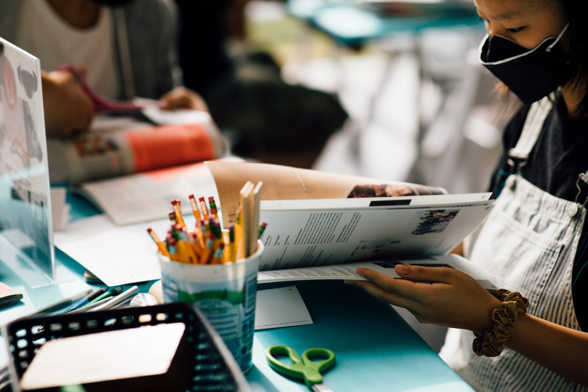 A person wearing a mask reads a magazine at a table with pencils, scissors, and paper.