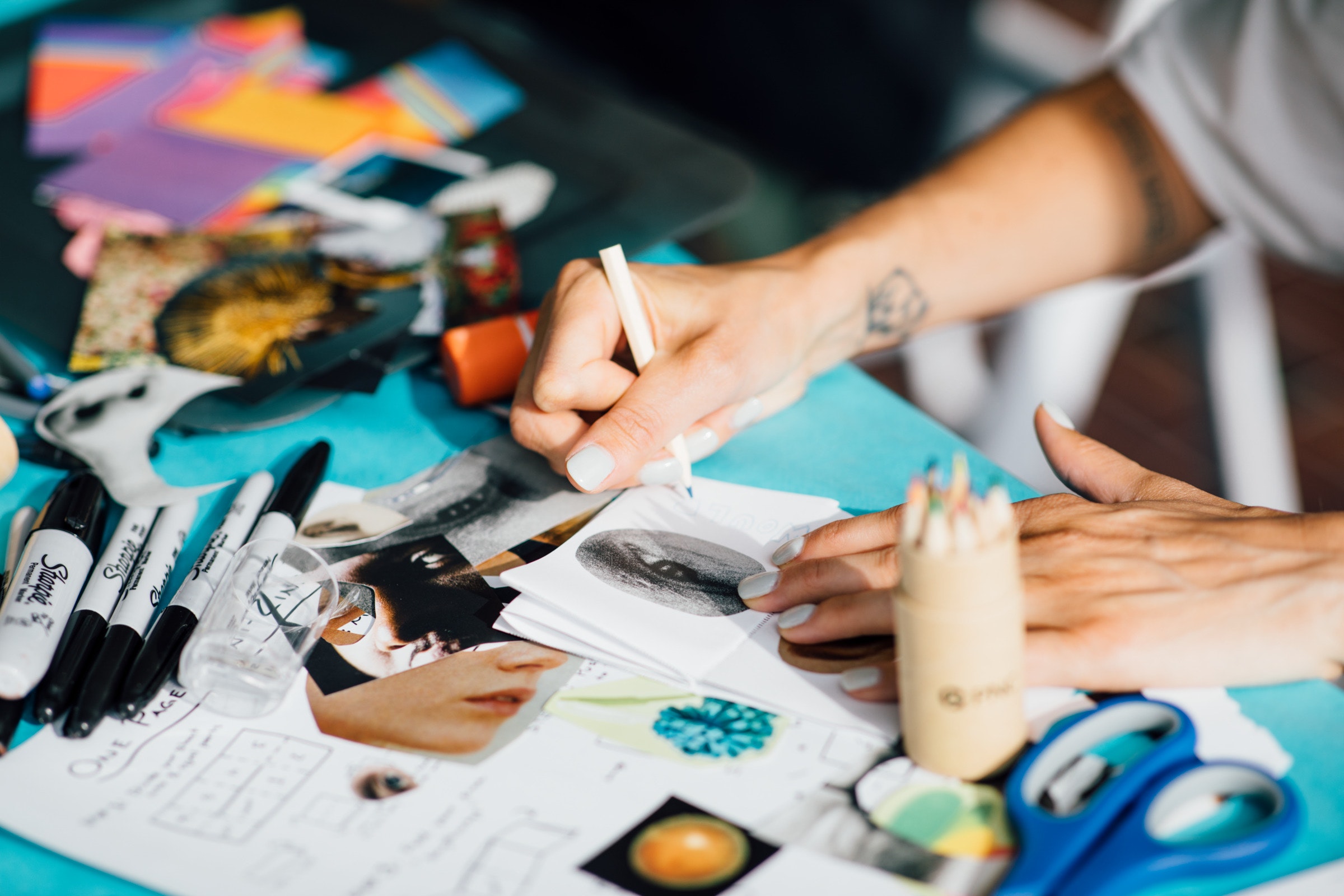 Person drawing on paper at a desk covered with colored pencils, markers, scissors, and collage materials.